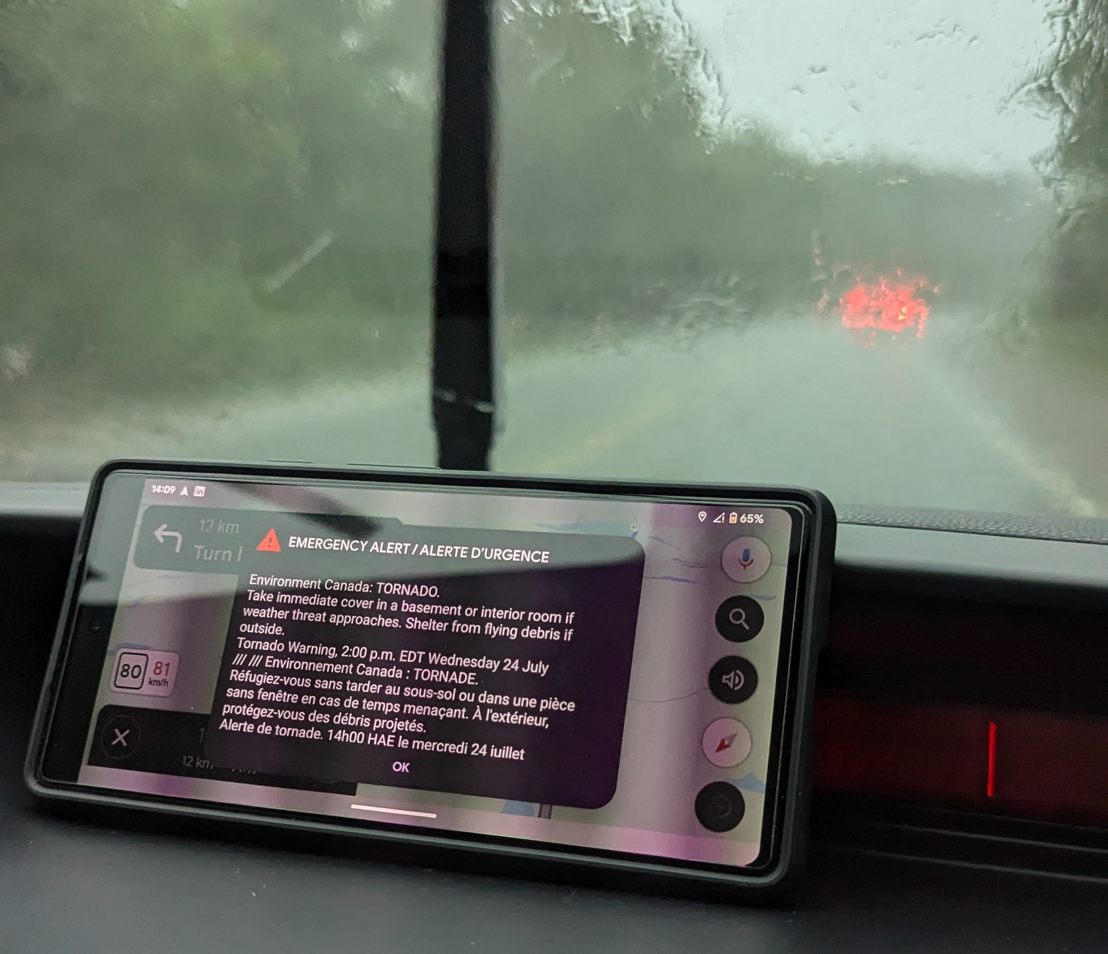 A phone on a car dashboard displaying an emergency alert for a tornado warning, with rain pouring on the windshield