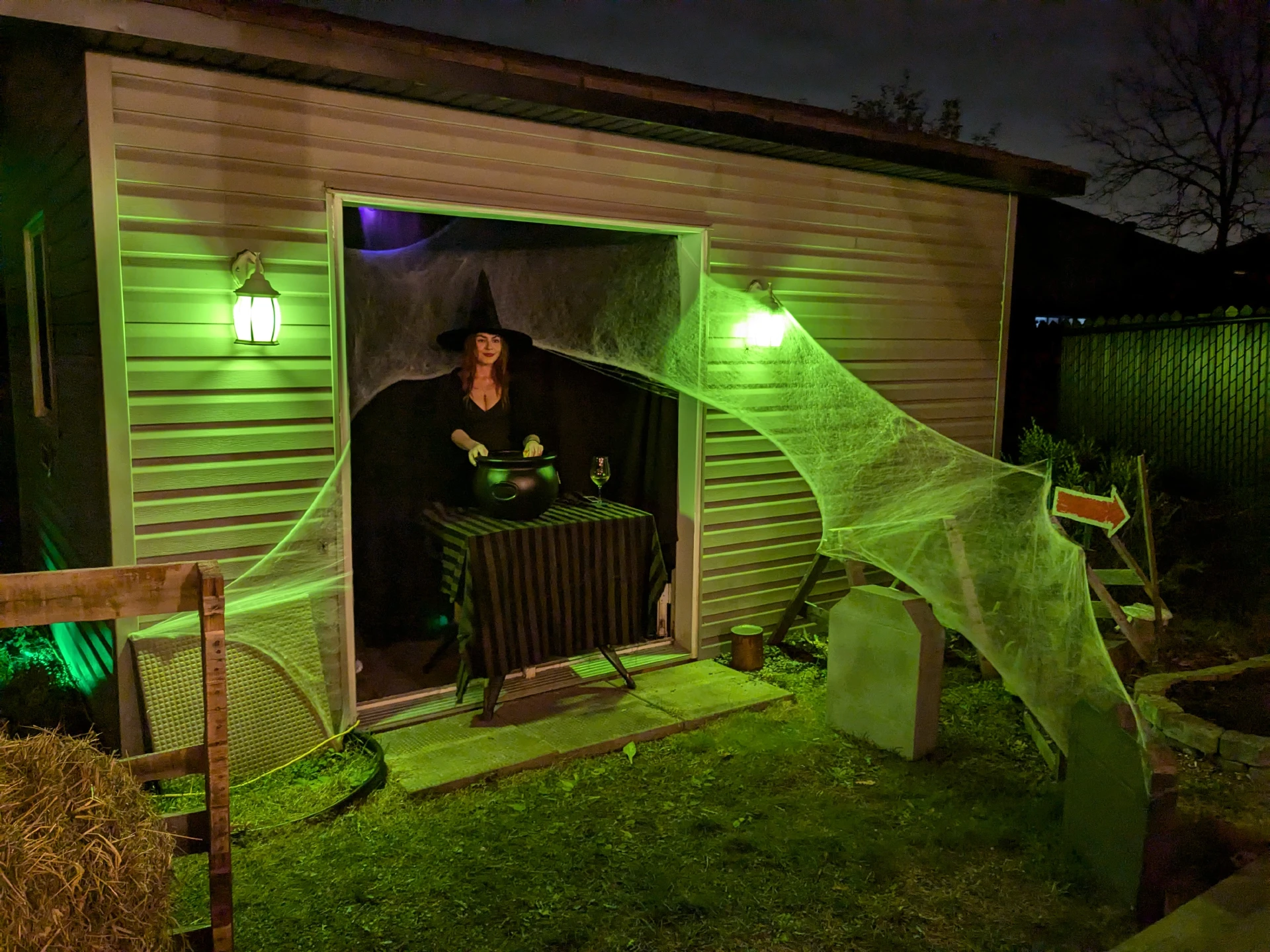 L standing with the candy cauldron, flanked by cobwebs bathed in green light from the shed lanterns